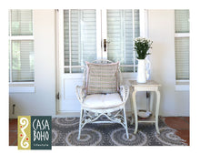 Load image into Gallery viewer, White wicker chair with a cushion in a room with windows and a small table.
